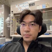Headshot of Gary Shi sitting at a desk chair with laboratory equipment in background