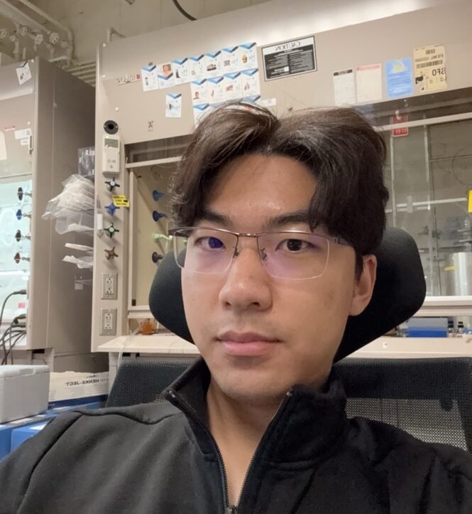 Headshot of Gary Shi sitting at a desk chair with laboratory equipment in background