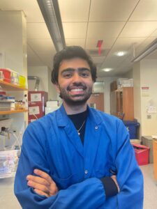 Riaz Mowzoon-Mogharrabi in a blue lab coat standing in front of a lab workstation